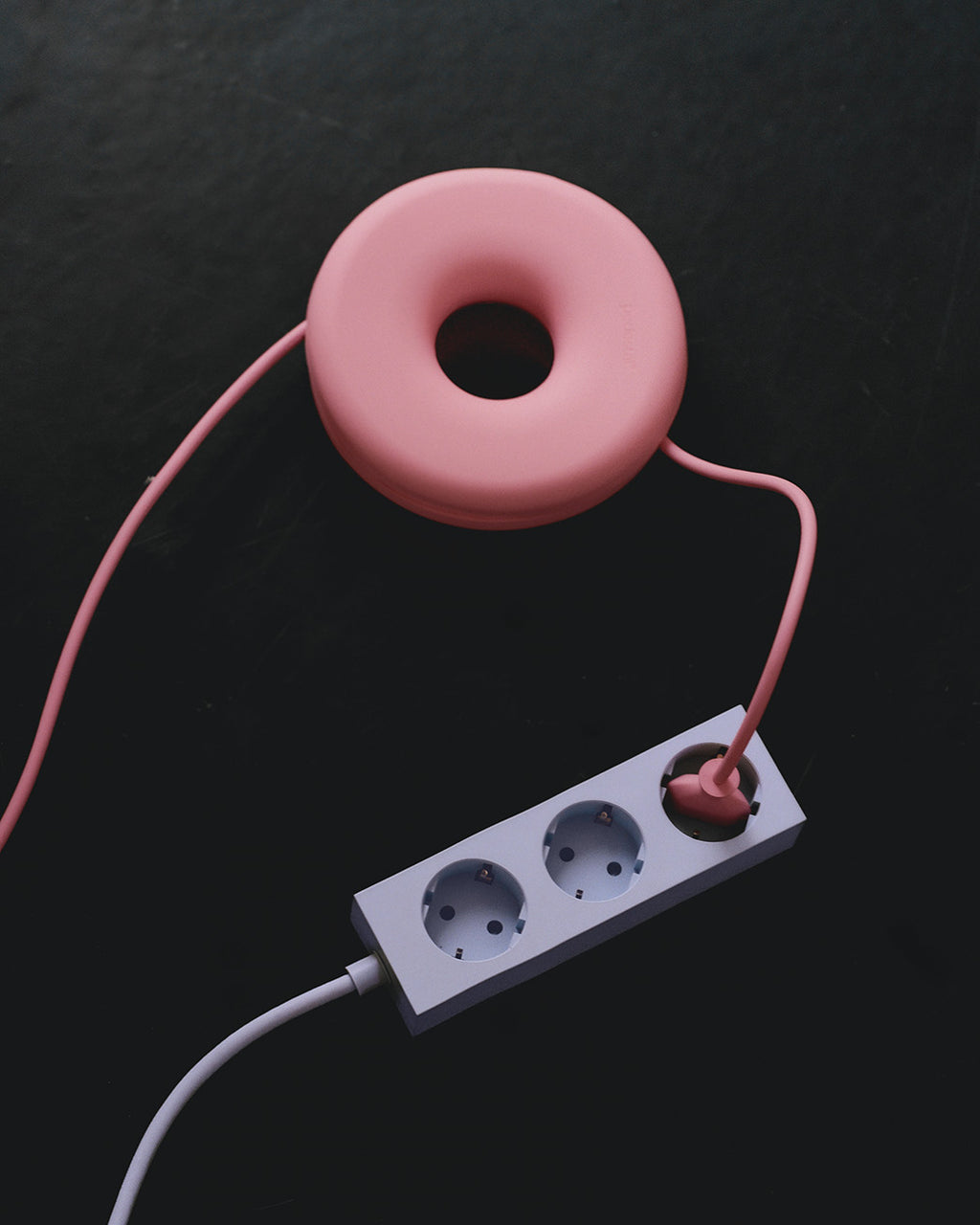 Bubble Gum pink Cable Bagel cable manager coiling a cord beside a white power strip on a dark textured surface