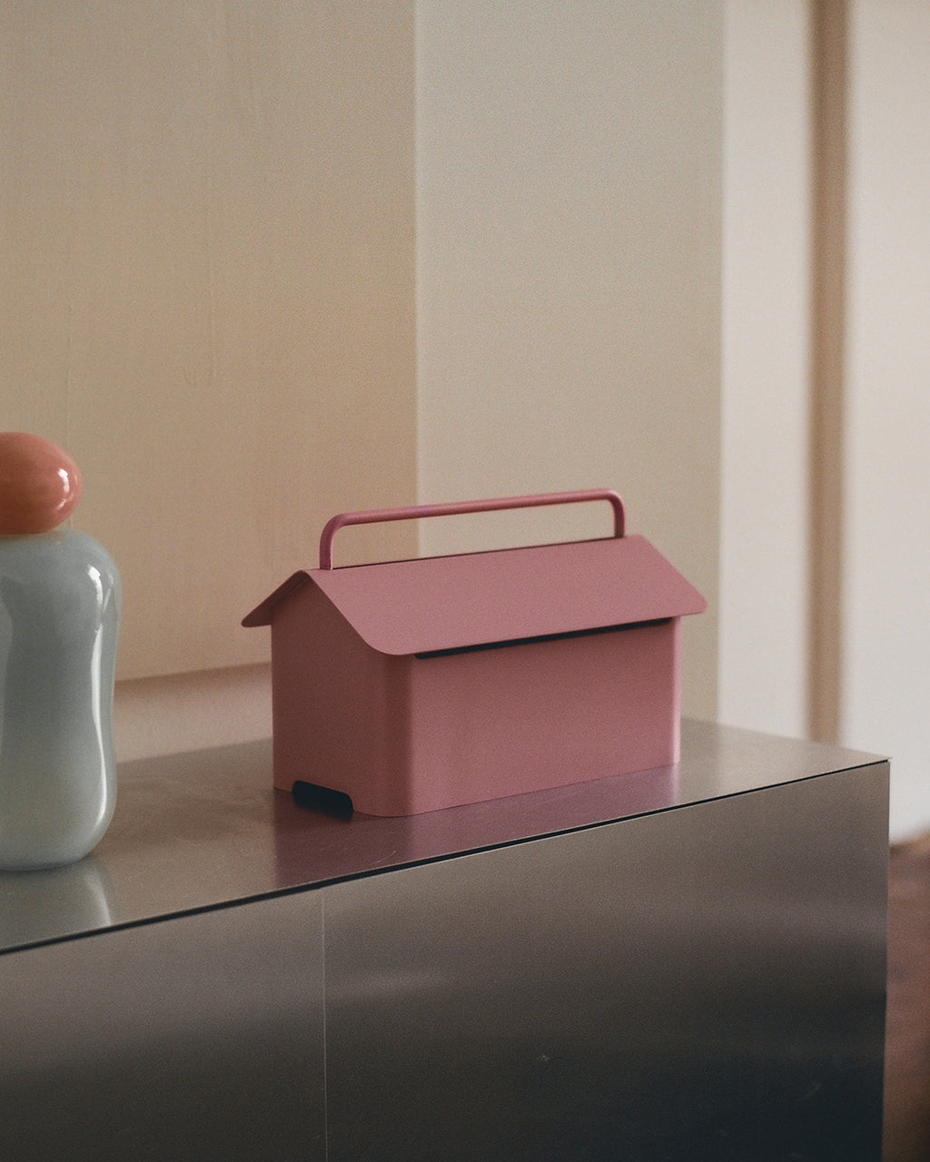 Cable Villa in Bubble Gum sitting on a steel console beside a pale vase, house-shaped lid and handle in view