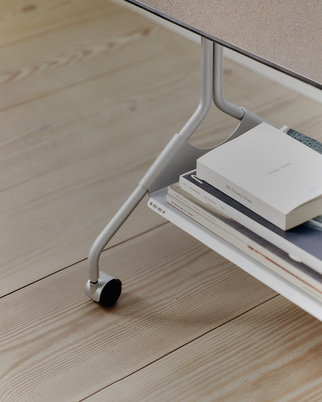 Close-up of Moon in Cloudy Silver stand's wheeled leg and lower shelf holding books on pale wood flooring