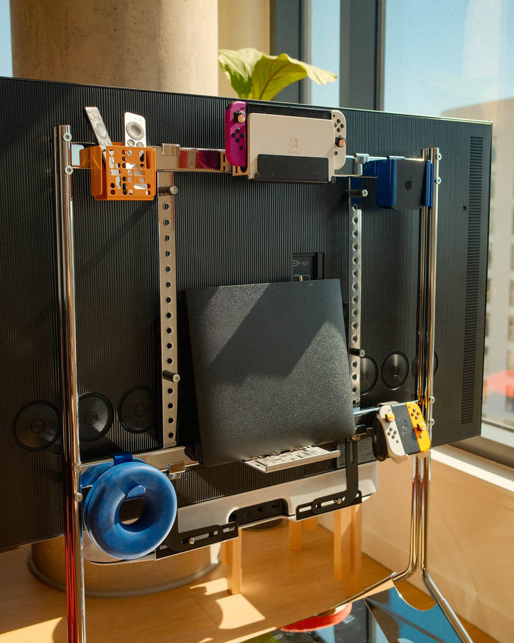Pearl Nintendo Switch Mount holding a Switch OLED behind a wall-mounted TV, alongside remotes and Joy-Cons on a rack